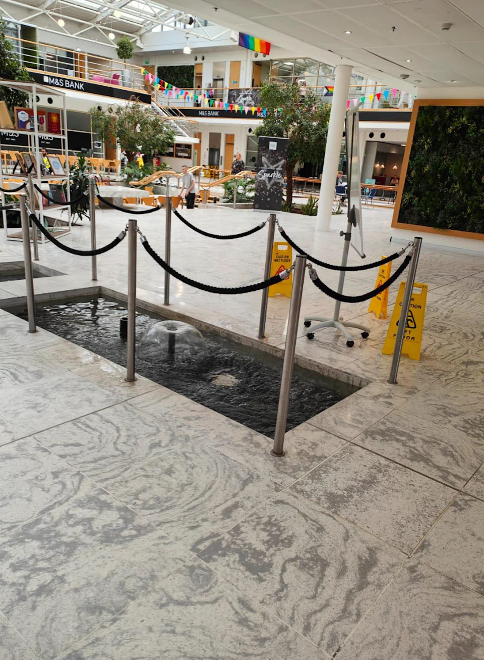 Inner mall scene shared with a narrow fountain pool divided by railing, damp floor tag nearby and shopping areas in the background