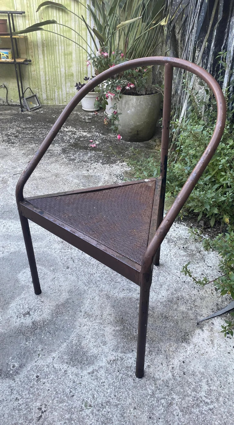 Metal chair with a minimalist, triangular seating design, which is surrounded on an outside terrace of plants