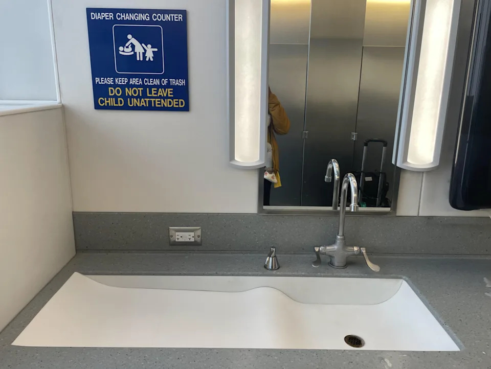Public bathroom with a diaper change station, shield that is pointed out not to leave a child unattended to leave a sink and a hand soap dispenser