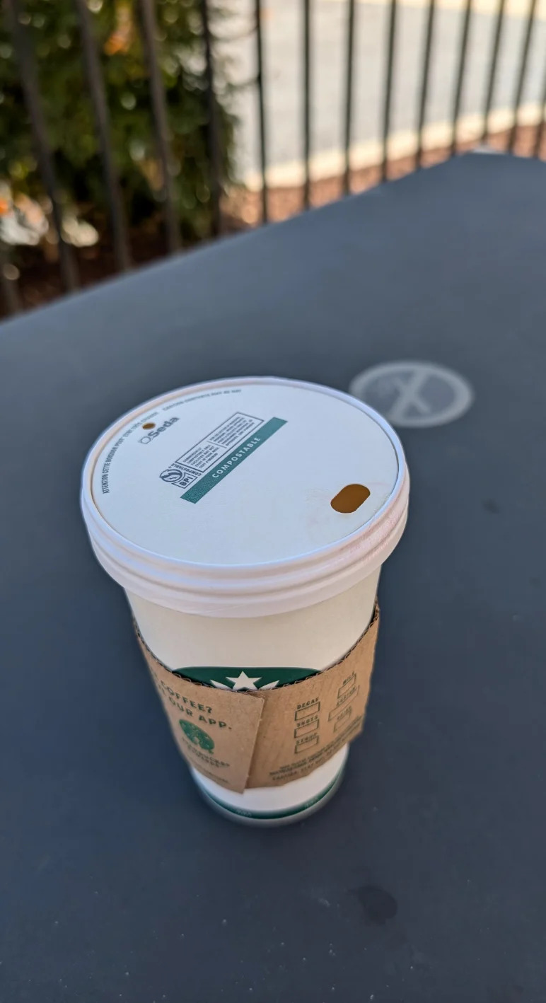 A coffee cup to take away with a secure lid that sits on a table outdoors near a metal fence