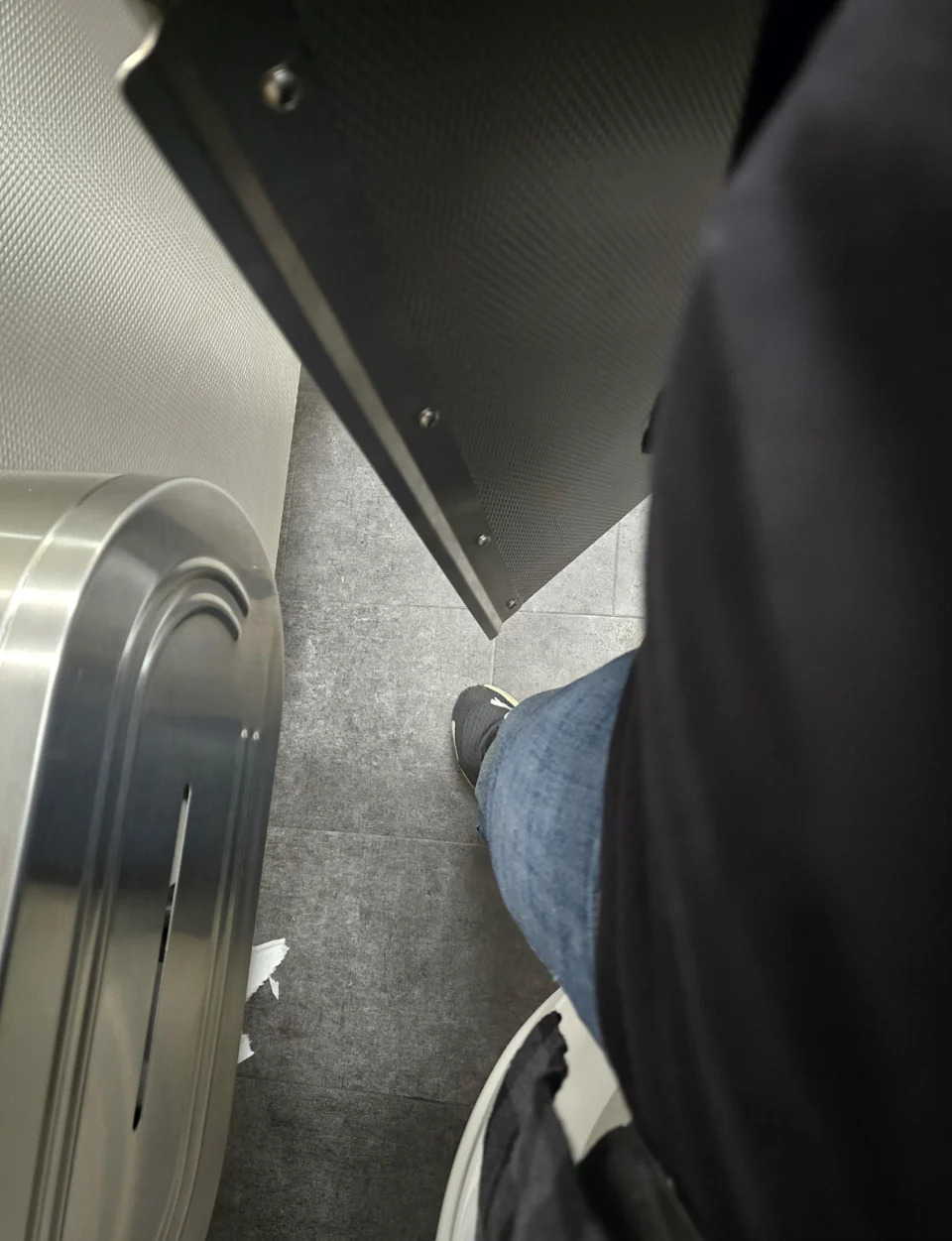 Person in a toilet stand, partly visible, with a leg in jeans and sneakers. Door slightly Ajar, which shows part of a metal container
