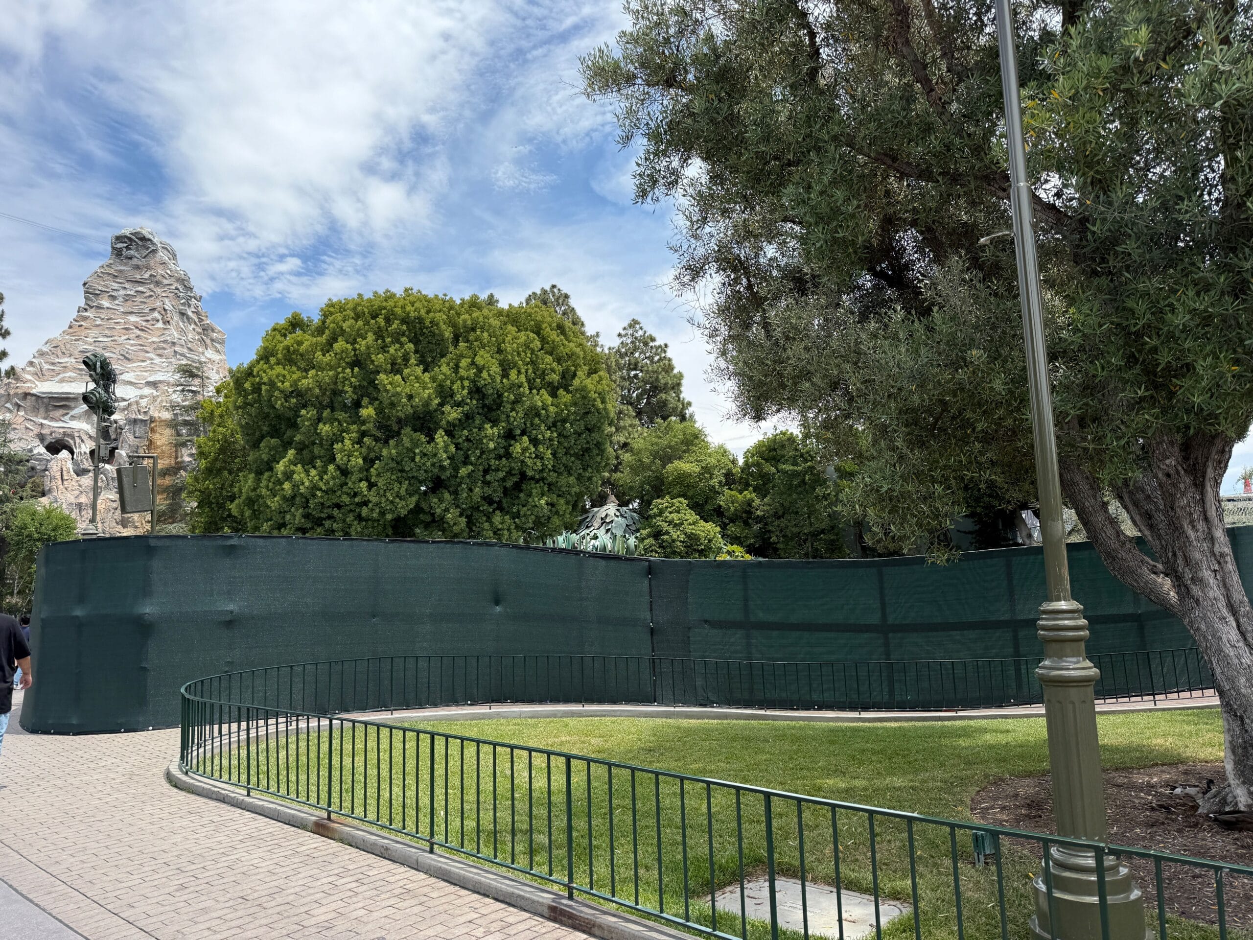 Green construction fences surrounds part of trees and lawn near a mountain construction in a parking environment.