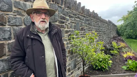 BBC John Watkeys stands on his new rock garden wall, which is approximately head height. He wears a tickling hat and glasses with a black coat and has a white beard. A flower bed can be seen on the right of the wall and grass on the right.