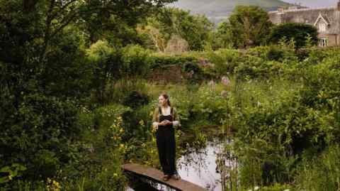 A woman in dungarees stands on a narrow wooden bridge over a pond, surrounded by lush green landscape