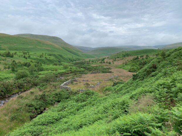 The landscape shot down a valley, with rolling green hills on both sides and in the distance. The sky is dark and cloudy. There are some stone walls in the foreground and a small river on the left of the picture.