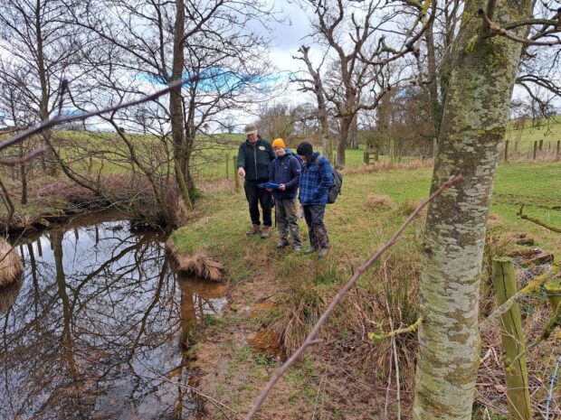 Three people in hiking equipment stood on the banks of a river and looked at an iPad. A man points to the river.