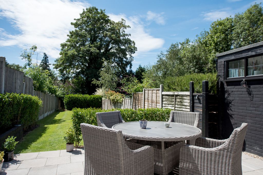 A general view that looks at a round garden with a round gray rattan garden table and chairs on the terrace on a sunny day with a blue sky and white clouds