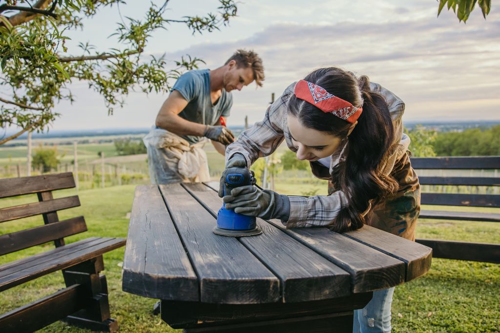 Adult couple, man with short brown hair and beard, woman with long brown hair, apron, grind her wooden table in the garden, woman with a blue grinding machine in the foreground, face near the machine while her husband paints the place