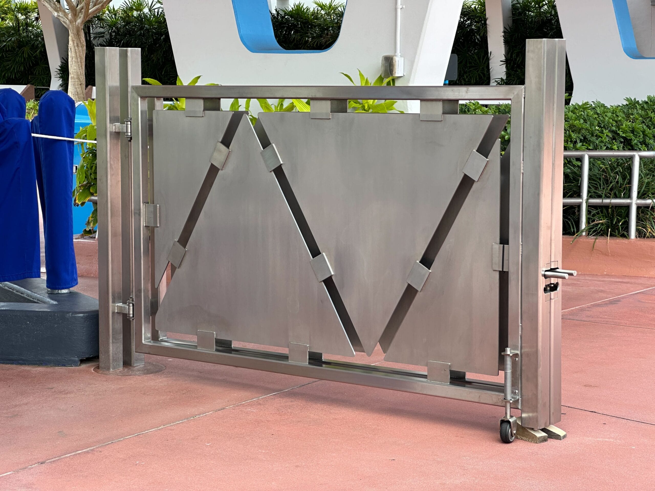 Modern metallic gate with geometric triangular patterns that are installed on a red sidewalk with green in the background.