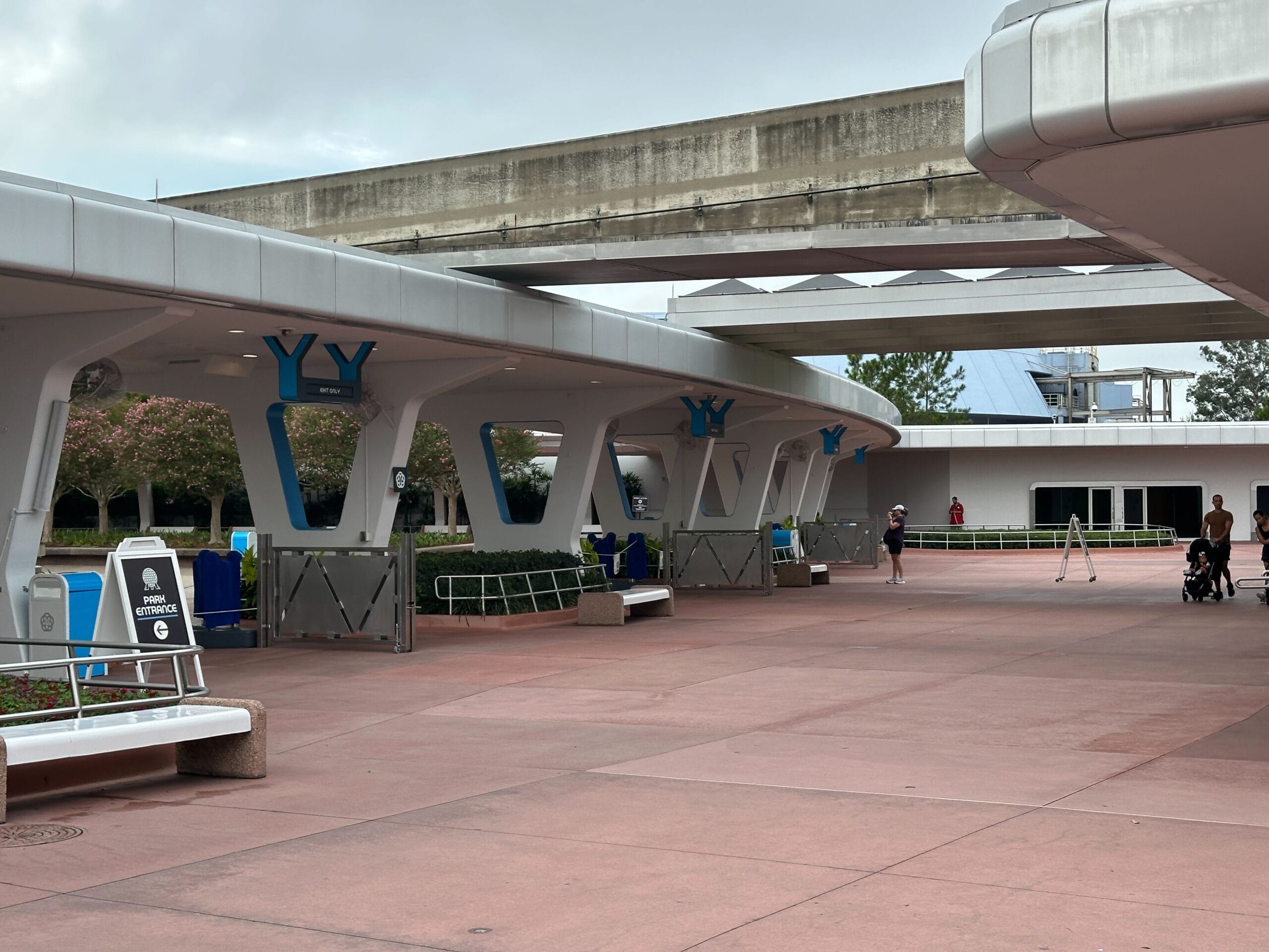 The curved sidewalk with white supports, blue accents and a monorail routing over the head on a mostly empty outdoor place.