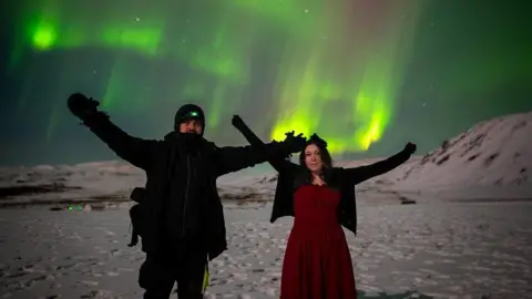 The Astro dog Simon Scott and Nicole Carr are in a snow -capped landscape and keep their arms with the green northern lights above them.