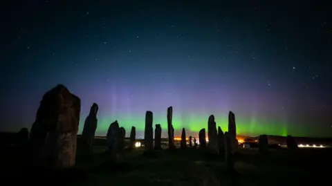 Josh Drury Callanish Stone Circle on the Isle of Lewis and The Northern Lights, a picture of the Somerset photographer Josh Drury, shows standing stones with a green and purple aurora behind and stars in the night sky.
