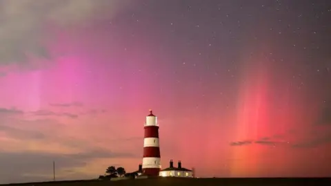 James Rowley-Hill James Rowley-Hills Photo by Pink, Red and Orange Northern Lights shows the Aurora over a red and white lighthouse. The cottage next to the lighthouse has its lights on.