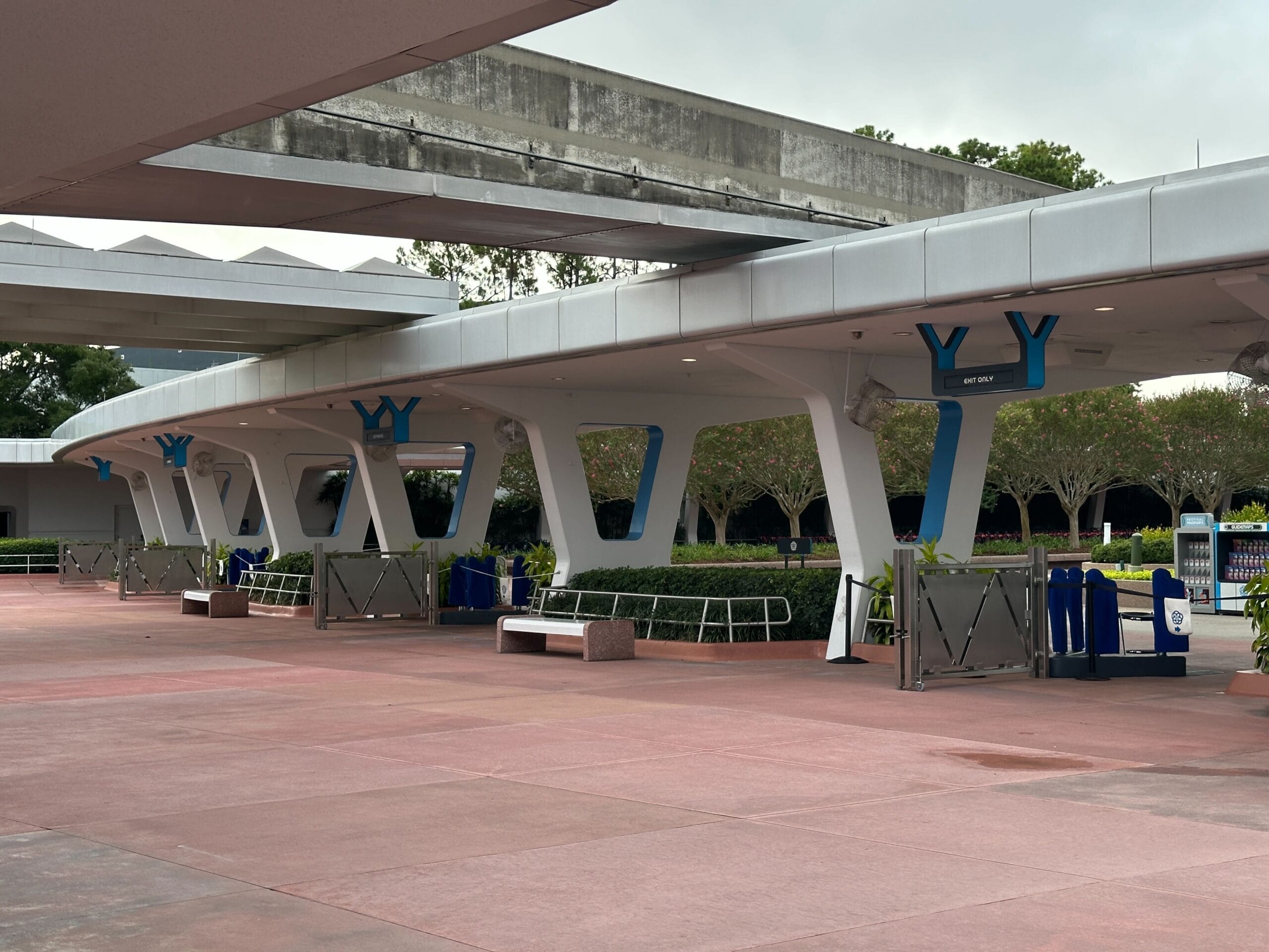 An almost empty walkway with modern white columns and blue signs with some benches and green in the background.
