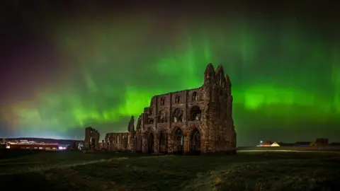 Astro Dog the Northern Lights in green and with red over Whitby Abbey in North Yorkshire. The brightest parts look like tapes in the sky. The ruins of the abbey stand with fields around them. There is a house in the background.