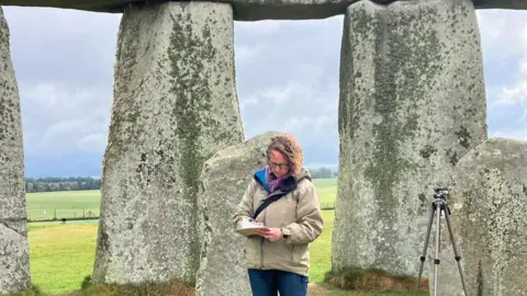 Jennifer Wexler Rose Ferraby wrote down some notes while studying Stonehenge. It stands in front of several standing stones, but lies in a circle and not outside the circle. The clouds are a moody gray and the sunlight breaks on the floor.