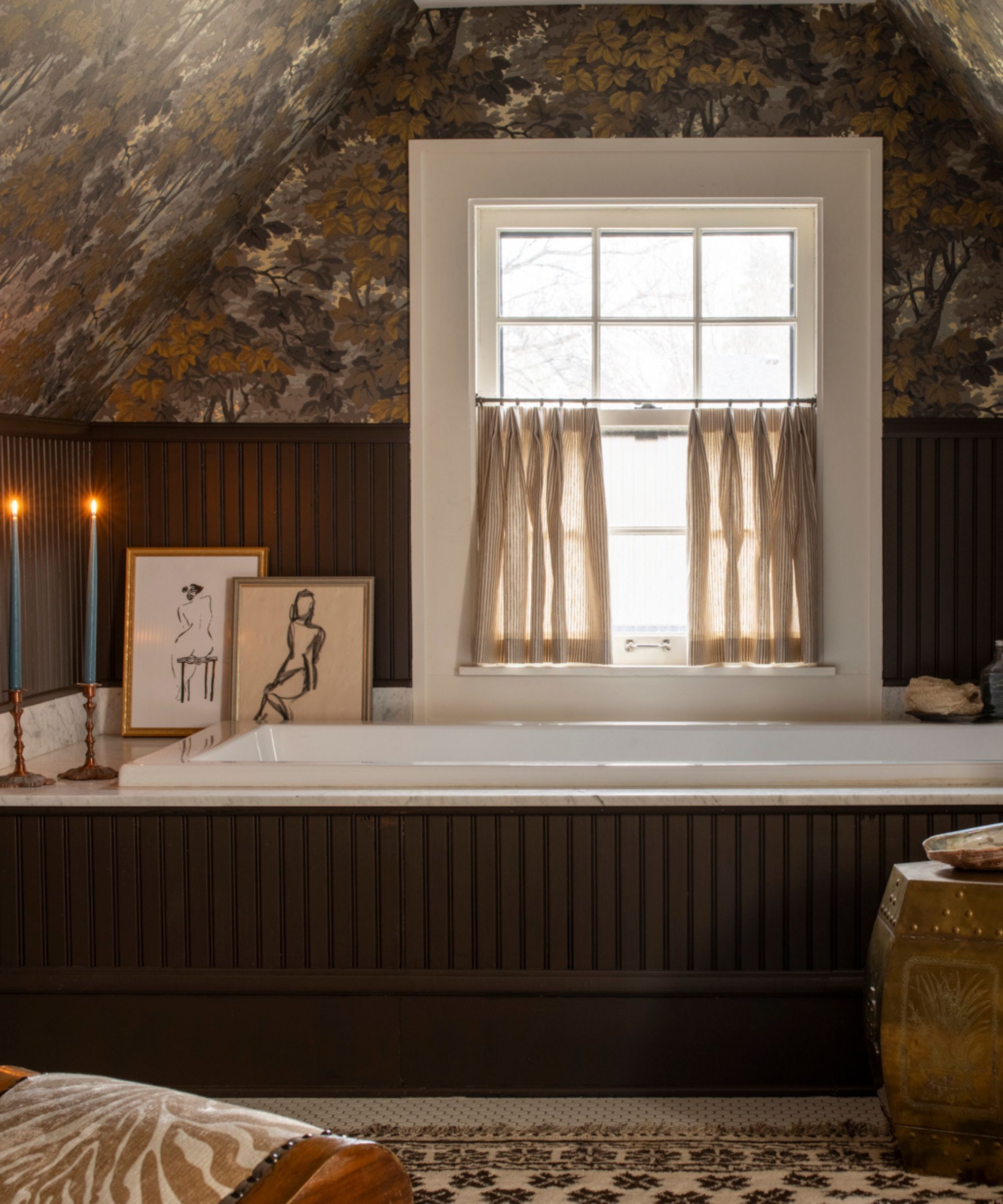 Atmospheric loft guest bathroom with dark brown wood paneling and bathtub surround, small window with cafe curtain, botanical wallpaper, decorated with a rug, stool, artwork, candles and a small chair