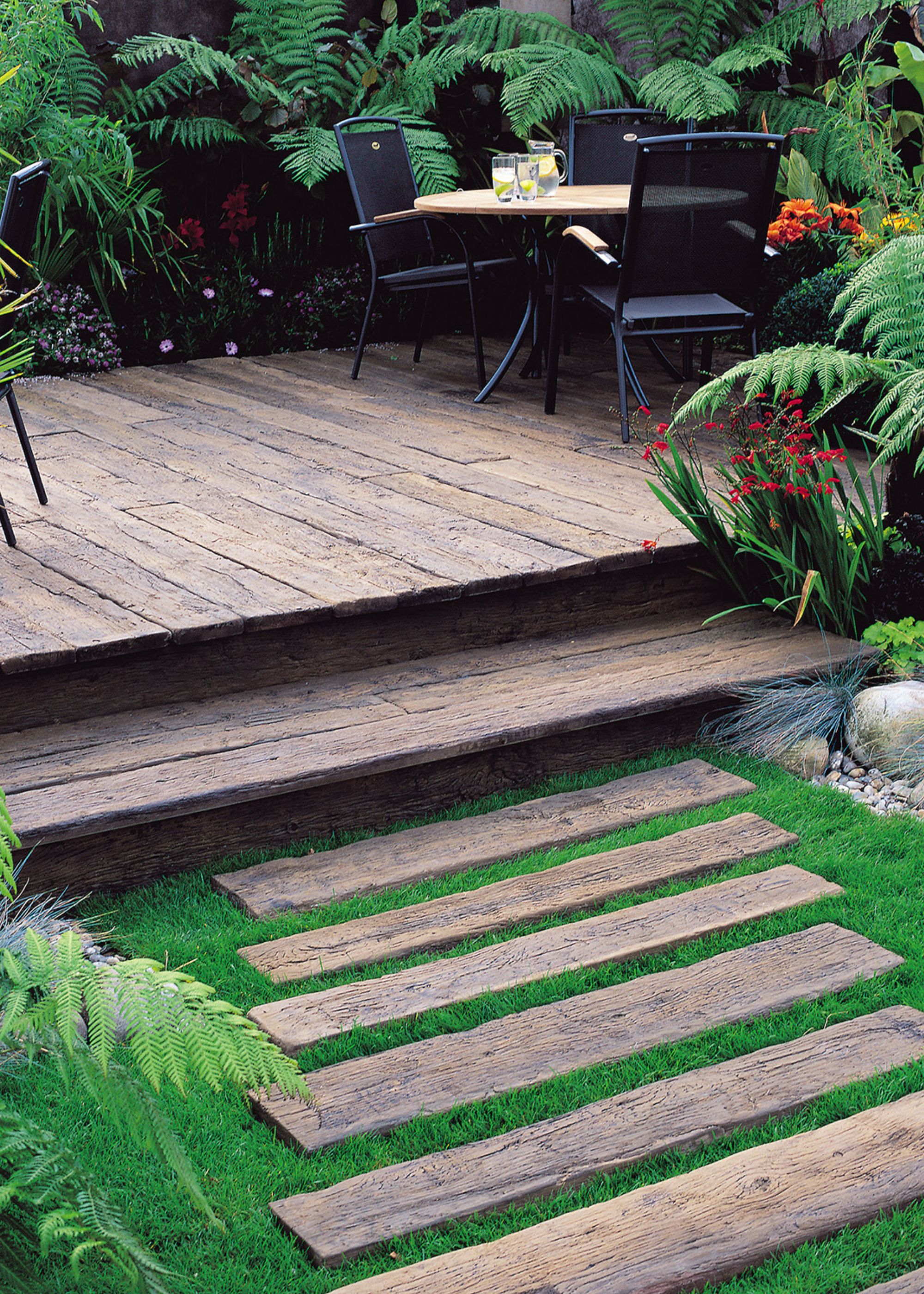 A garden with wooden steps and wooden terrace. There are plants and trees all around as well as a view of a dining table on the terrace and three chairs around it.