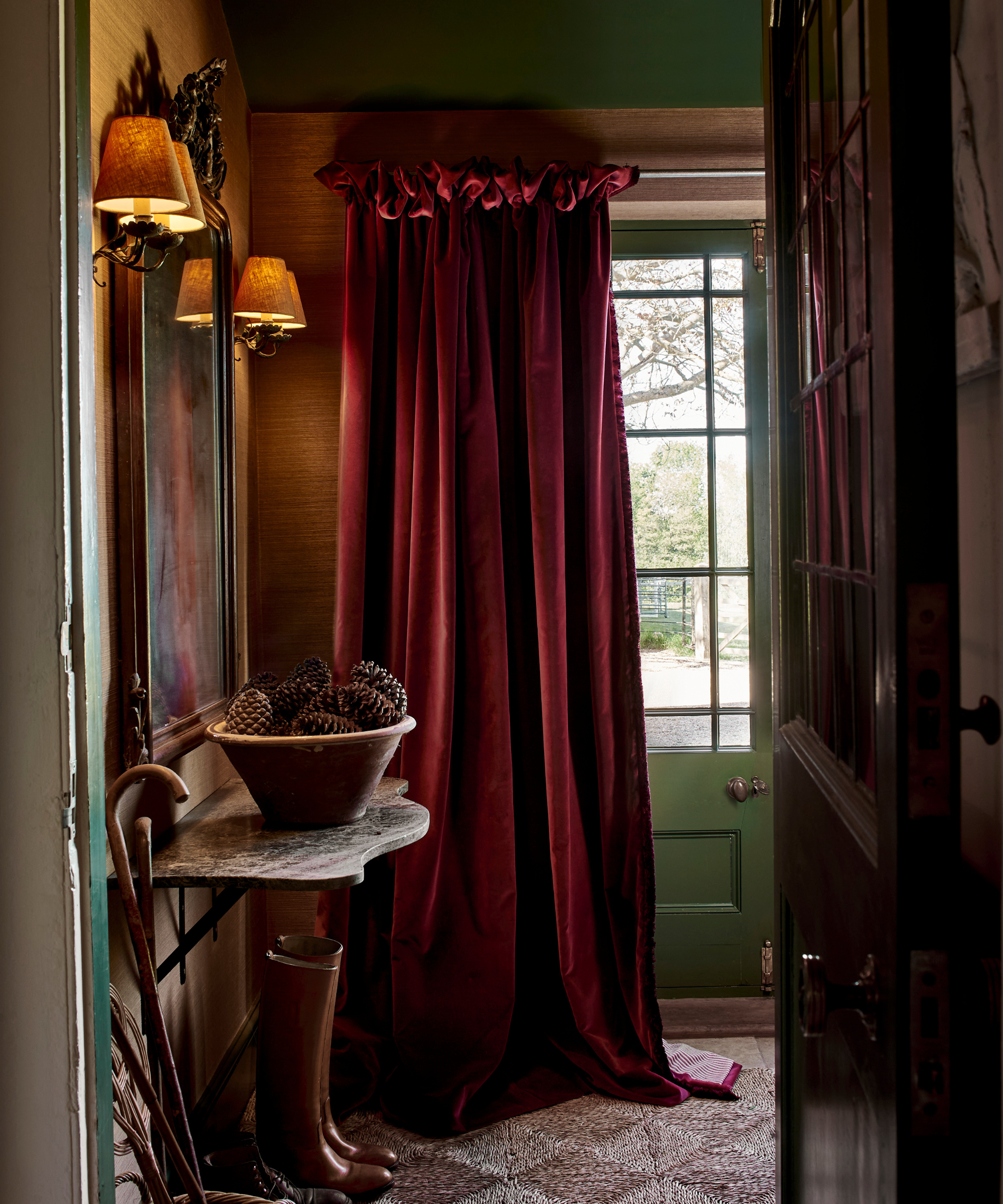 an entryway with a dramatic red velvet curtain, a marble table with a bowl of pine cones, a wall hanging artwork with two sconces on either side and a pair of boots on the floor