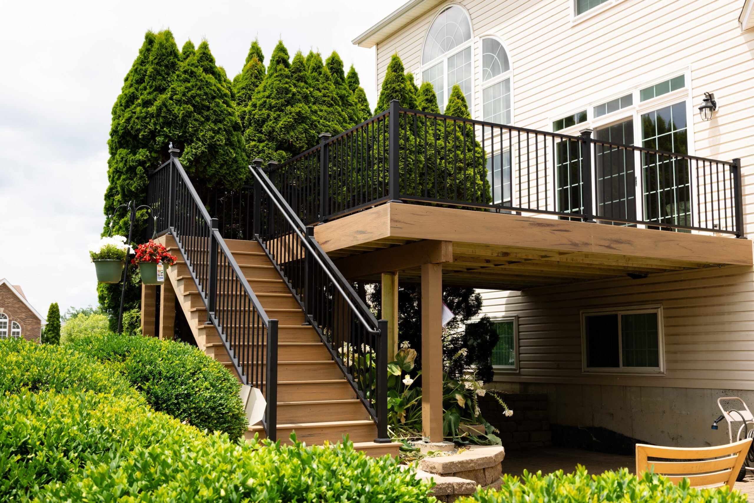Elevated backyard patio by Fence Deck and Depot with black metal railing and wooden stairs surrounded by lush green landscaping attached to a beige house with large windows