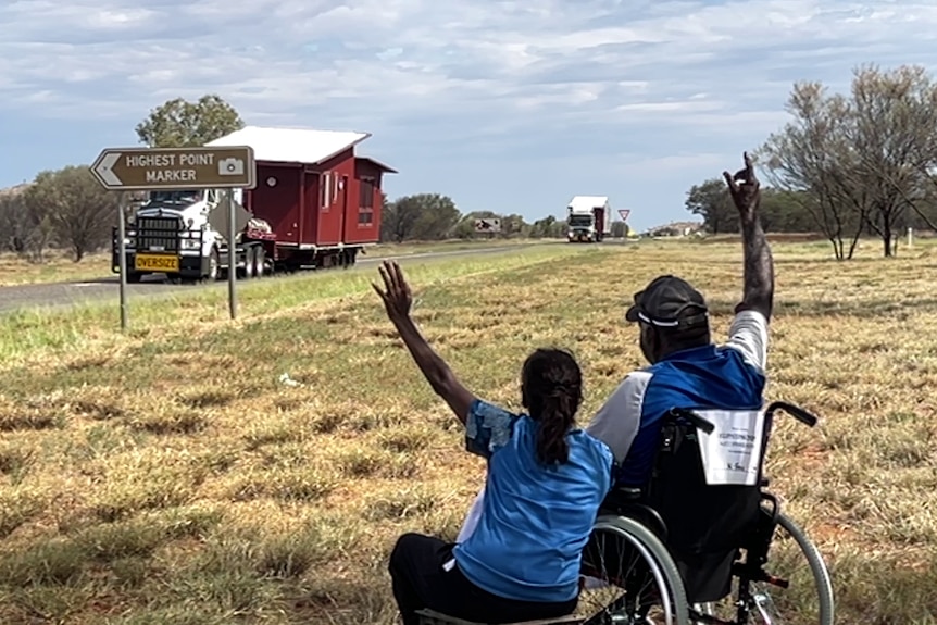 Two people sit in the grass on the side of a remote highway and wave to a passing truck carrying part of a house.
